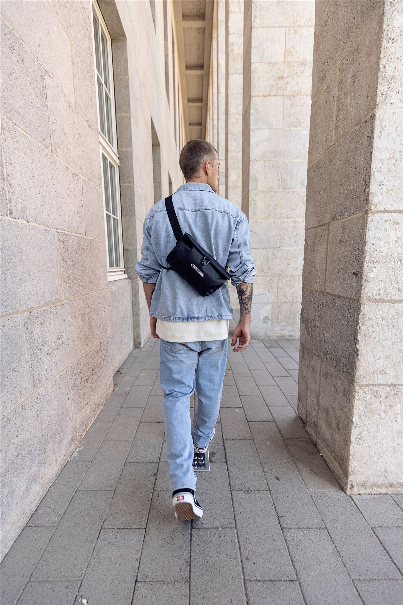 Rückansicht eines Mannes mit schwarzer ORTLIEB Velo-Sling Flex Umhängetasche mit Rollverschluss beim Gehen durch Säulengang.